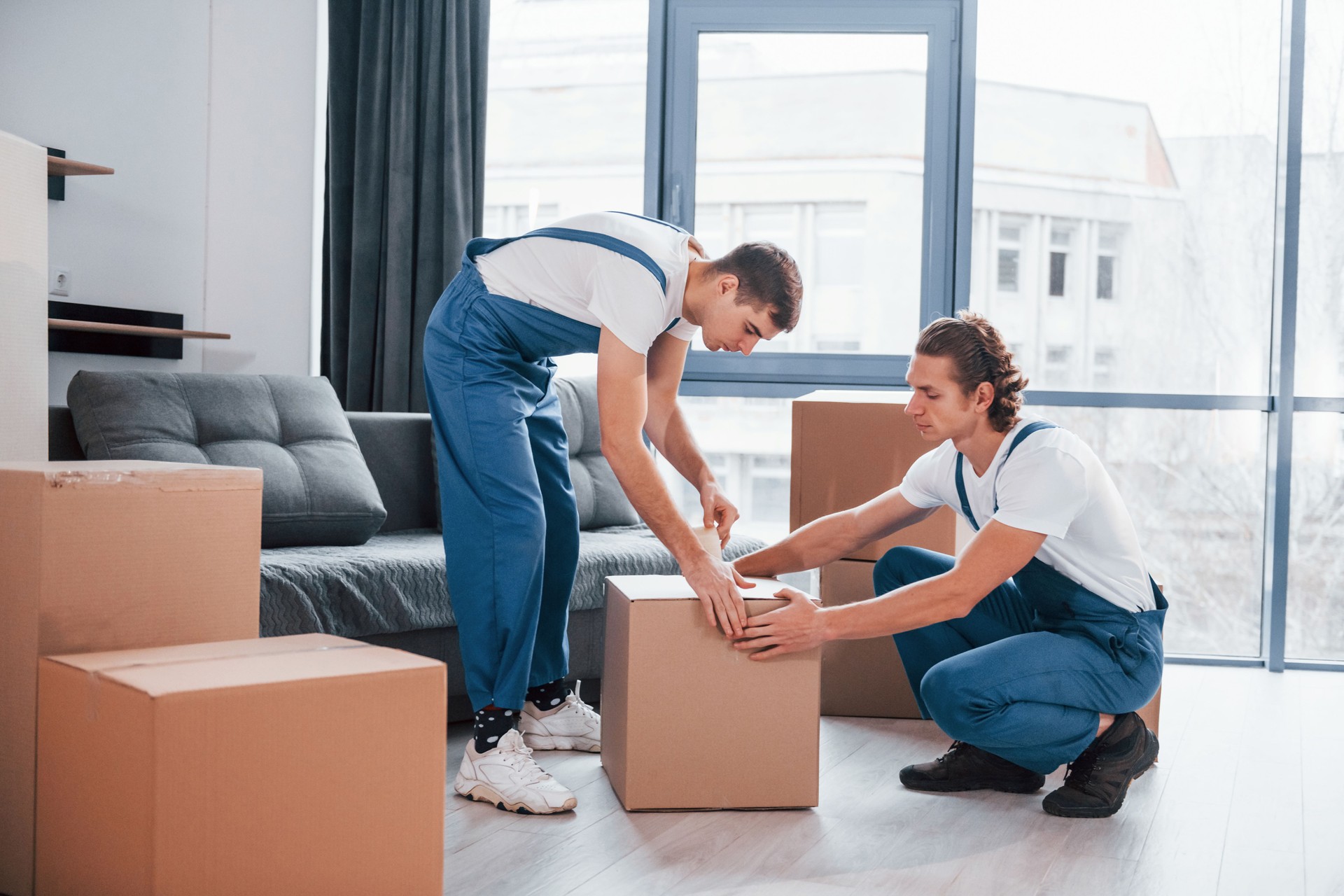 Packaging the box. Two young movers in blue uniform working indoors in the room